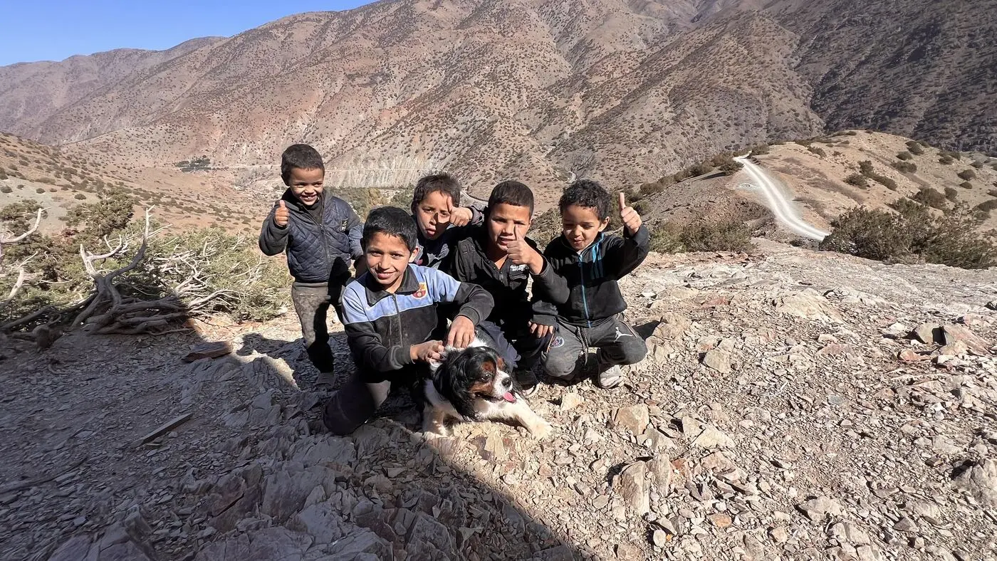 Five smiling boys give a thumbs-up beside Barclay, the School Aid mascot dog, high in Morocco's Atlas mountains