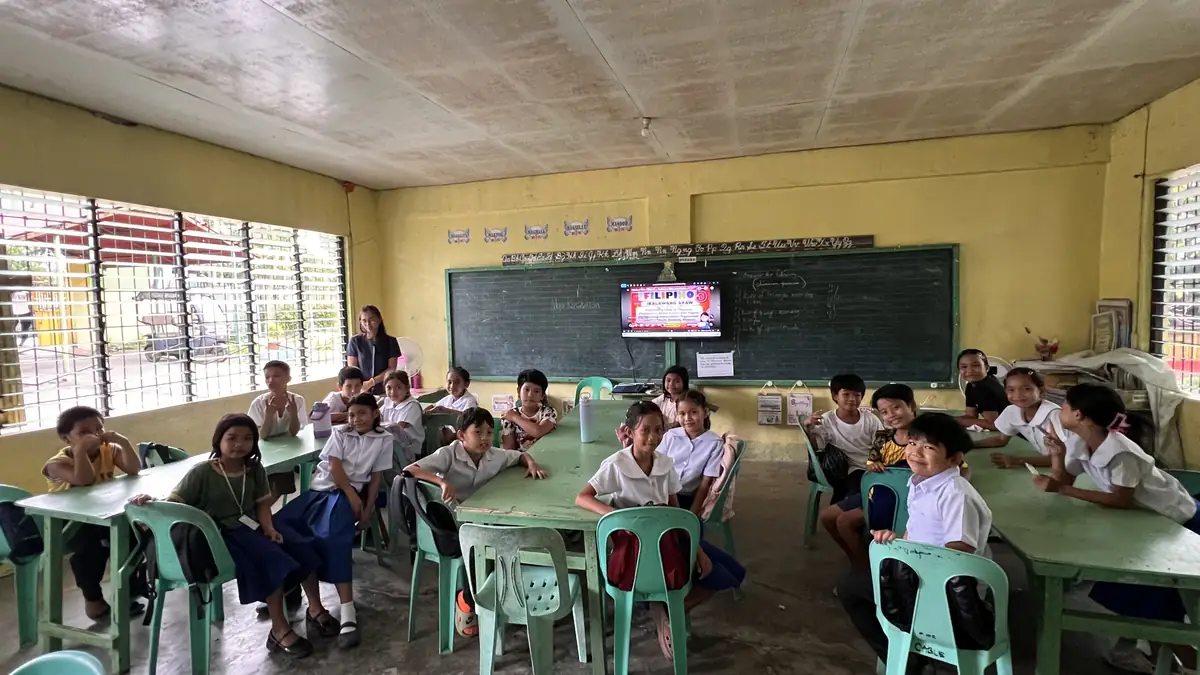 Pupils in school uniform seated at shared green tables in a classroom at Cabalabaguan Elementary School, with a single small screen at the front
