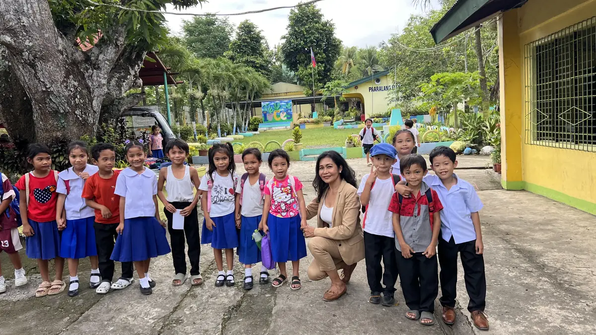 Maribeth de Montaigne of School Aid MTÜ crouching beside a line of pupils in school uniform on the grounds of Cabalabaguan Elementary School