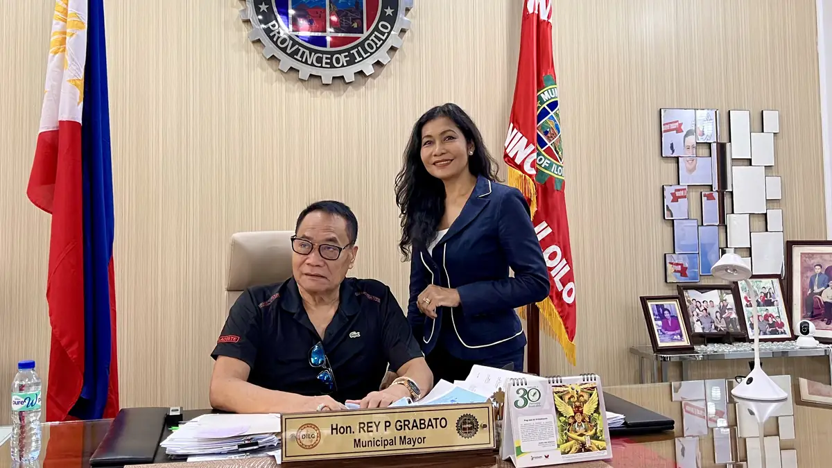 Mayor Rey P. Grabato of Mina, Iloilo, at his desk in the municipal office with Maribeth de Montaigne of School Aid MTÜ, beside the Philippine flag and the Mina municipal seal