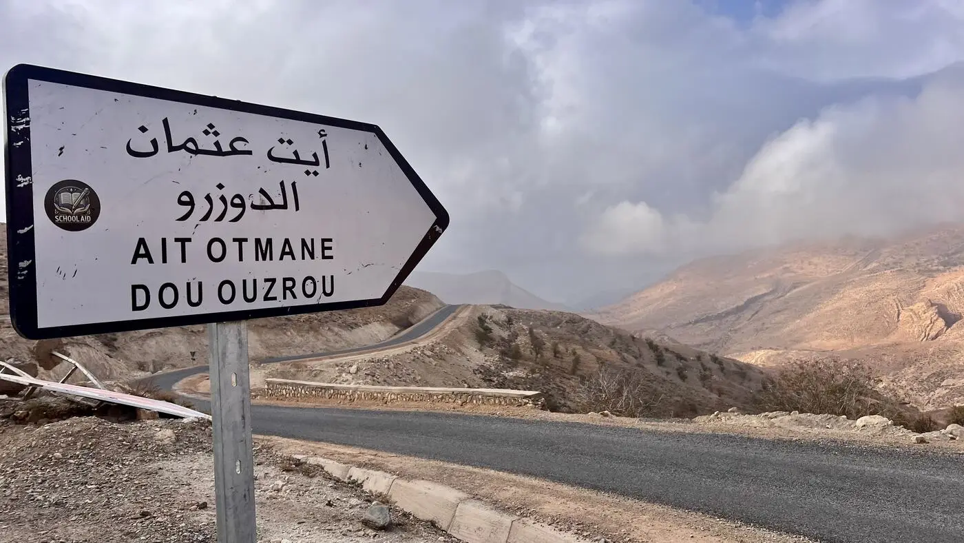 A mountain road sign for Ait Otmane bearing a School Aid badge, High Atlas